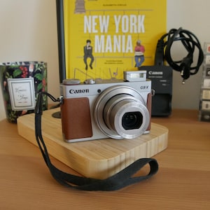 以下が含まれることがあります： シルバーとブラウンのCanon G9Xデジタルカメラで、黒いリストストラップが付いています。カメラには目立つレンズ、ポップアップ式フラッシュ、前面にCanonのロゴがあります。デバイスは明るい色の木製の表面に置かれています。