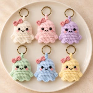 May include: Six crocheted ghost keychains arranged in two rows. Each ghost features a different pastel color, black bead eyes, a stitched smile, pink cheeks, and a small pink crocheted bow attached to the top. Each has a bronze-toned metal ring.