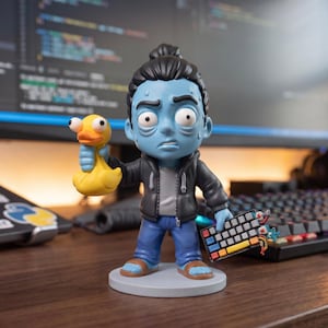 May include: A blue-skinned figurine with dark hair in a topknot, wearing a black hoodie, gray shirt, and blue jeans. The figure holds a yellow rubber duck in one hand and a small, multi-colored mechanical keyboard in the other, standing on a circular base.