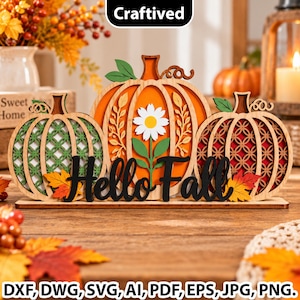 May include: A wooden tabletop decoration featuring three layered pumpkins with intricate cutouts, a central daisy, and the black script text Hello Fall. The left pumpkin is green, the center is orange, and the right is red, all on a base.