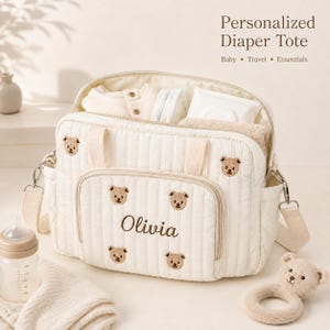 Könnte beinhalten: Eine cremefarbene, gesteppte Wickeltasche mit gestickten braunen Bärengesichtern und dem Namen Olivia in brauner Schreibschrift. Die Tasche verfügt über einen Reißverschluss, Seitentaschen und einen abnehmbaren Schultergurt, gefüllt mit Babyartikeln.