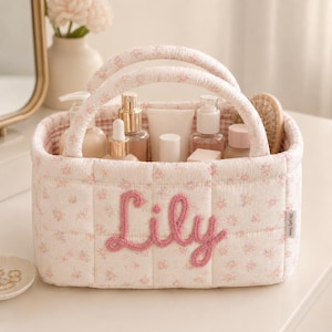 May include: A quilted fabric storage basket featuring a soft pink floral pattern on a white background. The front is embroidered with the name Lily in pink yarn. It has two rounded handles and contains various skincare bottles and containers.