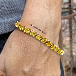 以下が含まれることがあります： 正方形にカットされた鮮やかな黄色の石が金属製のバンドにセットされたテニスブレスレット。ブレスレットの中央付近に「Labdiamondtreasure」という文字が見えます。均一なファセットカットの石が連結されたデザインです。