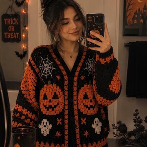 Könnte beinhalten: Eine schwarz-orangefarbene Häkelstrickjacke mit einem wiederkehrenden Muster aus Kürbissen, weißen Spinnennetzen und kleinen weißen Geistern. Das Kleidungsstück hat einen V-Ausschnitt, eine durchgehende Knopfleiste und langärmelige Strickbündchen.
