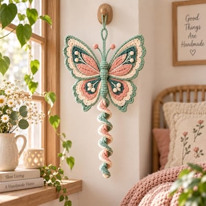 Könnte beinhalten: Ein gehäkelter Schmetterling als Wandbehang mit einem Körper in Salbeigrün, Flügeln in Salbeigrün, Altrosa und Creme sowie einem langen, spiralförmigen Schwanz aus abwechselnd salbeigrünen und altrosa Garnen, aufgehängt an einem Holzhaken.