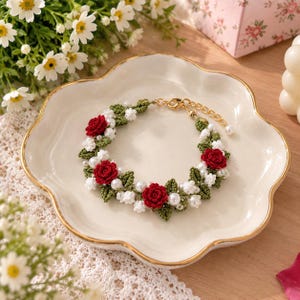 May include: A floral bracelet featuring crocheted red roses, small white flowers, and green leaves interspersed with white pearls. The bracelet includes a gold-toned metal chain with a lobster clasp closure and an adjustable extender link.