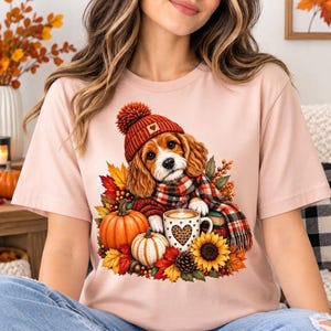 Puede incluir: Una camiseta rosa claro con el gráfico de un perro spaniel marrón y blanco que lleva un gorro de punto rojo y una bufanda de cuadros. El perro está rodeado de calabazas naranjas, hojas de otoño, un girasol y una taza con un corazón de leopardo.