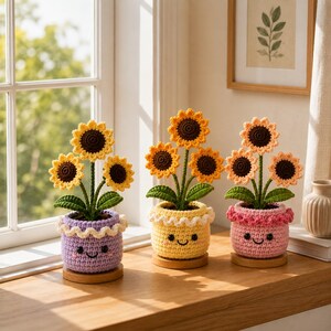 Könnte beinhalten: Drei gehäkelte Sonnenblumenpflanzen in kleinen, runden, lächelnden Töpfen. Der linke Topf ist lavendelfarben, der mittlere gelb und der rechte rosa. Jede Pflanze hat drei Sonnenblumen mit dunkelbraunen Mitten und gelben oder pfirsichfarbenen Blütenblättern.