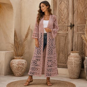 Könnte beinhalten: Ein langer, vorne offener Cardigan in Altrosa mit einem komplizierten, durchgehenden Häkelspitzenmuster mit Blumenmotiven, langen, ausgestellten Ärmeln und einem geraden Saum, der bis zur Mitte der Wade reicht, getragen über einem weißen Oberteil und Jeans.