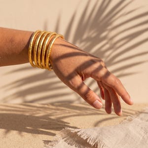 May include: A set of five gold-coloured bangles with a textured, matte finish. The bracelets are circular, thin, and stacked together on the wrist, featuring a consistent metallic yellow hue and a slightly uneven, organic surface texture.