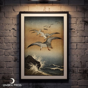 May include: A framed wall art print featuring a Japanese-style illustration of three seagulls flying over crashing ocean waves against a gradient tan and blue sky. The artwork is enclosed in a simple black frame and displayed on a brick wall.