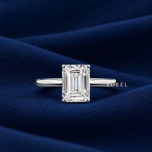 May include: A silver-toned ring featuring a large, rectangular emerald-cut center stone with faceted edges. The band is smooth and polished, showcasing a clean, reflective surface. The text ZORÉL is visible in small white lettering near the ring base.