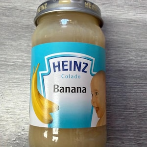May include: A glass jar of Heinz banana baby food with a silver metal lid. The blue and white label features the Heinz logo, the text 'Colado' and 'Banana', an illustration of two yellow bananas, and a graphic of a smiling baby's face.