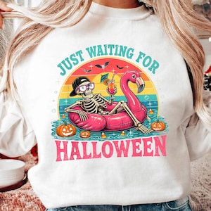 May include: A white sweatshirt featuring a graphic of a skeleton wearing a black baseball cap and sunglasses, lounging in a pink flamingo pool float. The design includes the text JUST WAITING FOR HALLOWEEN, pumpkins, bats, and a cocktail.