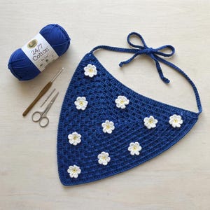 Puede incluir: Un pañuelo triangular de ganchillo azul oscuro con aplicaciones de flores blancas y centros amarillos. El conjunto incluye un ovillo de hilo 24/7 Cotton, un ganchillo de madera y unas tijeras plateadas, dispuestos para un proyecto.