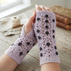 May include: A pair of light purple fingerless gloves featuring an intricate crochet pattern with small, dark black floral embellishments scattered across the surface. The gloves have a ribbed cuff at the wrist and an opening for the thumb.