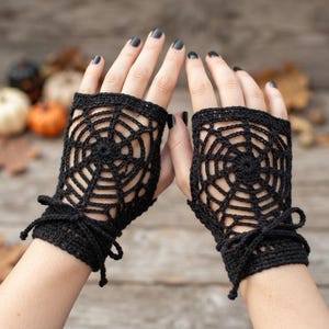 May include: A pair of black fingerless gloves featuring a crocheted spider web pattern. The gloves are constructed with a circular web design covering the back of the hand and include a small decorative tie at the wrist for a secure fit.
