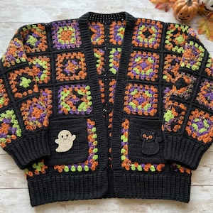 May include: A long-sleeved crochet cardigan made of black, orange, purple, and lime green granny squares. The garment features a solid black ribbed trim, a V-neckline, and two front pockets. A beige ghost patch is on the left pocket and a black cat patch is on the right pocket.