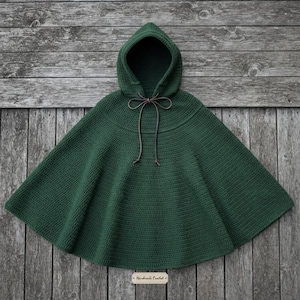 May include: A dark forest green hooded cape crafted with a crochet stitch pattern. The garment features a rounded hood and a thin brown leather cord tie closure at the neckline. A small rectangular wooden tag labeled Handmade Crochet is attached.
