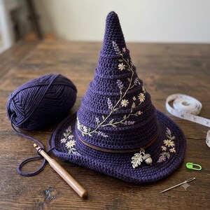 May include: A dark purple crocheted pointed hat featuring embroidered floral vines, a brown leather band, and silver-toned leaf and acorn charms. The hat is accompanied by a ball of matching purple yarn, a wooden crochet hook, a needle, and a stitch marker.