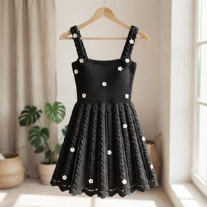 Könnte beinhalten: Ein ärmelloses Häkelkleid in Schwarz mit quadratischem Ausschnitt, dicken geflochtenen Trägern und einem plissierten Rock mit Zopfmuster. Das Kleid ist mit kleinen, weißen, fünfblättrigen Blumenapplikationen verziert, die über das Oberteil und den Rock verteilt sind.