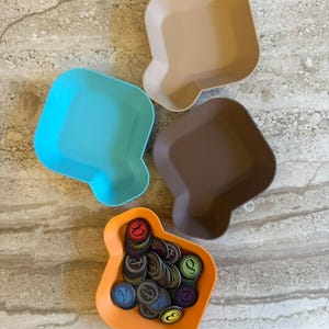 Op de afbeelding: Vier vierkante plastic schaaltjes met afgeronde hoeken en een kleine schenktuit aan één kant. De schaaltjes zijn lichtbeige, fel turquoise, donkerbruin en oranje van kleur. Het oranje schaaltje bevat een verzameling kleine, ronde speelfiches.