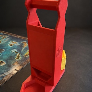 Puede incluir: Una torre de dados vertical roja con un asa en la parte superior y una bandeja base hexagonal. El lateral de la torre presenta un logotipo en relieve con el texto HeroQuest. Un pequeño recipiente amarillo está unido al lateral de la estructura.