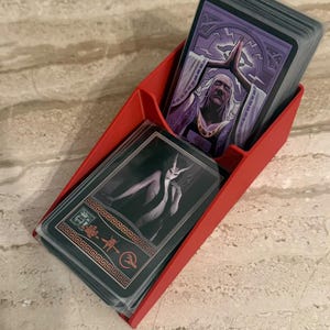 May include: A red plastic two-tier card holder containing two decks of playing cards. The top deck features an illustration of a person with white hair in a prayer pose, while the bottom deck shows a dark figure with horns and red symbols.