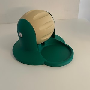May include: A dark green plastic base with a circular tray and a rotating beige spherical component featuring three horizontal indentations. The device has a mechanical hinge on the side and a smooth, matte finish throughout its structure.