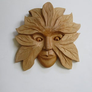 May include: A wooden carving of a face with leaves surrounding it. The face is carved in a realistic style with detailed features. The leaves are carved in a stylized way and are arranged around the face in a symmetrical pattern.