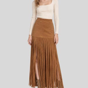 May include: A long, tan suede skirt featuring a high-waisted design and a floor-length hemline covered in long, vertical fringe detailing. The skirt includes two prominent side slits that reveal the legs, paired with a cream-coloured ribbed top.