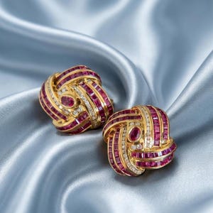 Könnte beinhalten: Ein Paar goldene Ohrringe in Knotenform, besetzt mit zahlreichen quadratisch geschliffenen roten Rubinen und kleinen runden weißen Diamanten. Jeder Ohrring ist mit einem oval geschliffenen Rubin in der Mitte versehen, was ein dichtes Muster ergibt.