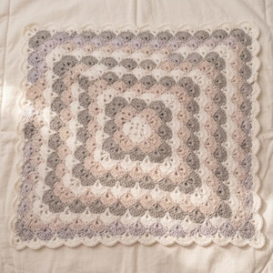 Può includere: Una coperta quadrata all'uncinetto con un motivo ripetuto a punto conchiglia in una palette di colori crema, rosa tenue, grigio chiaro e lavanda. La coperta è rifinita con un bordo smerlato color crema, creando un design strutturato.