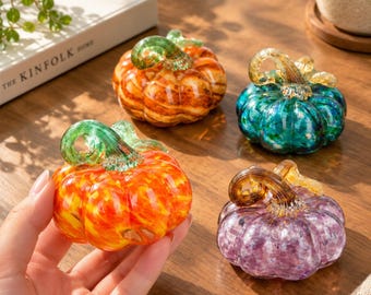 Könnte beinhalten: Vier kleine Glaskürbisse in Orange, Türkis, Gelborange und Violett. Jeder Kürbis weist eine ausgeprägte gerippte Textur und einen klaren, geschwungenen Glasstiel auf. Die Objekte sind auf einer Holzoberfläche angeordnet.