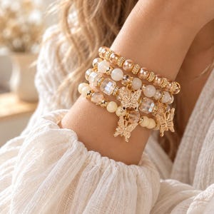 Peut inclure: Un empilement de quatre bracelets de perles composé d'un mélange de perles crème, champagne et transparentes à facettes. Les bracelets sont agrémentés d'entretoises dorées et de deux breloques papillon dorées suspendues.