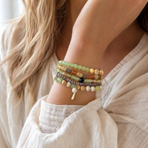 May include: A stack of four beaded bracelets featuring a mix of light green, amber, grey, and white beads. The bracelets include gold-toned metallic accents, a single black bead, and a small white stone charm shaped like a tapered cylinder.
