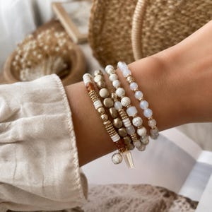 May include: A stack of four beaded bracelets featuring a mix of white marble-patterned beads, metallic gold-toned discs, small gold-toned spheres, translucent iridescent beads, and a single amber-coloured bead, finished with a small dangling charm.