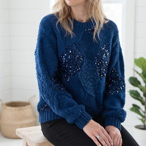 May include: A deep blue long-sleeve sweater featuring a textured knit pattern with alternating sections of cable knit and fuzzy yarn. The front of the sweater is embellished with dark blue sequins arranged in diamond shapes across the chest and sleeves.