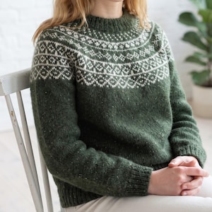 May include: A forest green long-sleeve jumper featuring a round neckline and a white geometric fair isle pattern across the yoke. The knit garment has a ribbed collar, cuffs, and hem, with subtle flecks of lighter colour throughout the wool fabric.