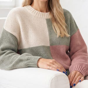 Puede incluir: Un suéter de punto de manga larga con un diseño de bloques de color con grandes paneles rectangulares en crema, verde salvia y rosa empolvado. La prenda tiene un cuello redondo acanalado y puños acanalados, confeccionada con una textura de punto grueso.