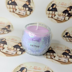 May include: A round glass candle holder with a crackled texture and an iridescent finish, filled with light purple wax and a single white wick. The front of the glass features the printed text Candle, Ice Pixie, and the brand name Aroma Alley.