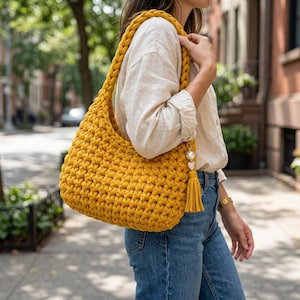 May include: A mustard yellow crochet shoulder bag featuring a thick braided strap and a textured knit pattern. The bag is accented with a decorative charm consisting of a white pearl bead, a small heart, and a matching yellow fabric tassel.