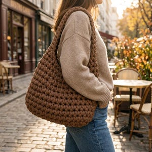 May include: A brown crochet shoulder bag featuring a thick, braided strap and a chunky, textured knit pattern. The bag is constructed from thick yarn loops, creating a soft, rounded silhouette that hangs from the shoulder.