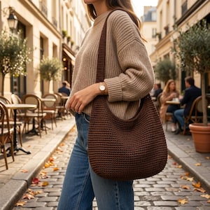 May include: A dark brown crochet hobo bag with a wide shoulder strap, worn with a beige ribbed knit sweater and blue denim jeans. The bag features a dense, textured stitch pattern and a curved, slouchy silhouette that hangs at the hip.
