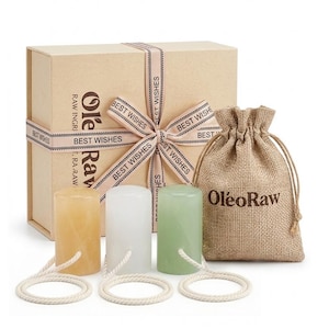 Könnte beinhalten: Eine beigefarbene Geschenkbox mit einem Band mit der Aufschrift BEST WISHES, ein beigefarbener Jutebeutel mit Kordelzug und dem Text OleoRaw sowie drei zylindrische Massagesteine in Gelb, Weiß und Grün, jeweils mit einer weißen Kordelschlaufe.