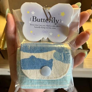 Könnte beinhalten: Ein kleiner Stoffbeutel mit blau-weißem Muster, der mit einem hellblauen Druckknopf verschlossen wird. Er ist in einer durchsichtigen Plastiktüte verpackt, mit einer schmetterlingsförmigen Karte, die den Text "Butterfly" und "Every time I see you, I feel so calm, like a butterfly flying over the ruins." enthält.
