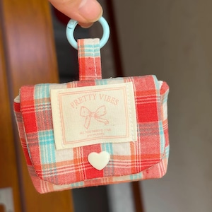 Könnte beinhalten: Ein kleines Stofftäschchen mit einem rot-weiß-türkisfarbenen Karomuster. Die Frontklappe hat ein cremefarbenes, rechteckiges Etikett mit dem Text "PRETTY VIBES" und einer Schleifengrafik sowie einen kleinen weißen Knopf in Herzform. Ein hellblauer Ring ist befestigt.