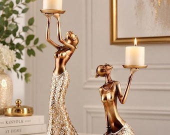May include: Two bronze-toned candle holders shaped like figures. One figure stands holding a white pillar candle overhead, while the other kneels holding a candle. Both figures feature textured, floral-patterned skirts in a metallic gold finish.