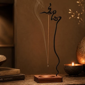 May include: A vertical incense holder featuring a dark metal wire frame with a calligraphic design at the top, mounted on a rectangular wooden base. A single incense stick is held upright, emitting a thin trail of smoke into the air.