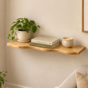 Peut inclure: Une étagère murale en bois clair avec un bord avant ondulé. Sur l'étagère se trouvent une plante verte dans un pot en céramique crème, deux livres empilés intitulés Live Beautifully et The Kinfolk Home, et un petit bougeoir rond couleur crème.